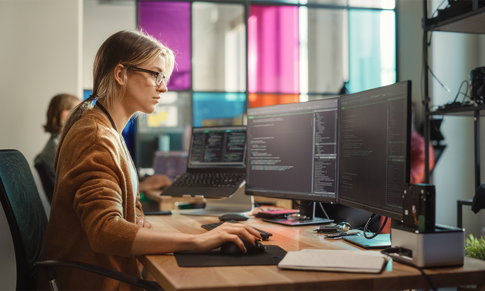 HR-Solutions_Frau-vor-mehreren-Bildschirmen Frau sitzt vor mehrereb. Bildschirmen an ihrem Arbeitsplatz, Ansicht von der Seite