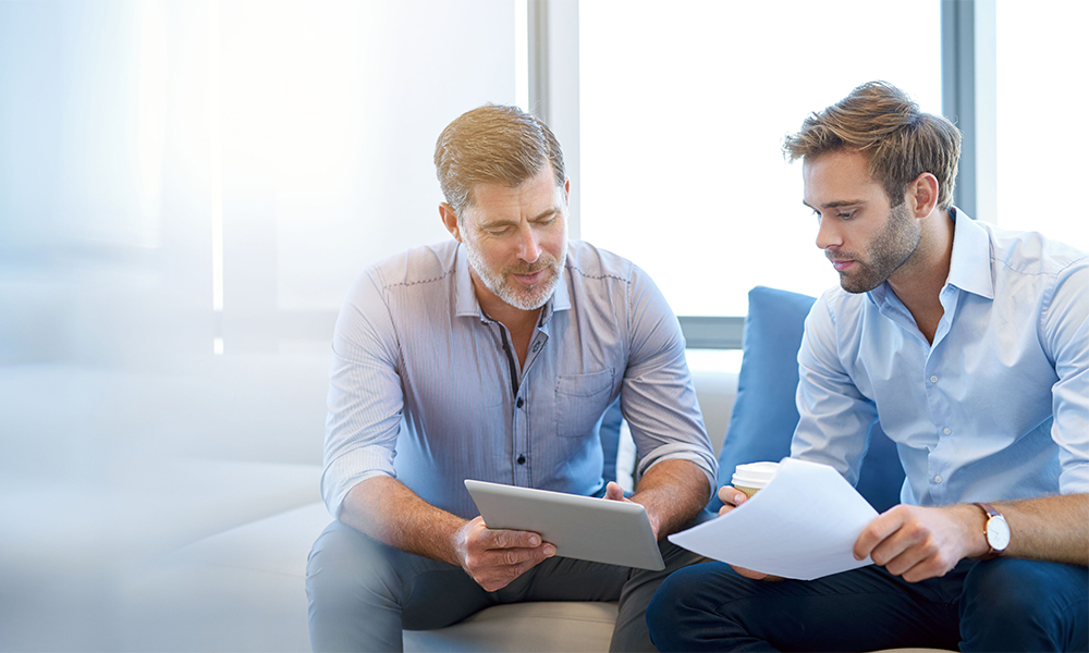 HR-Solutions_Kollegen-Unterhaltung-schauen-auf-Tablet Geschäftsmann im Gespräch mit jüngerem Kollegen auf Couch, dabei schauen sie gemeinsam auf ein Tablet