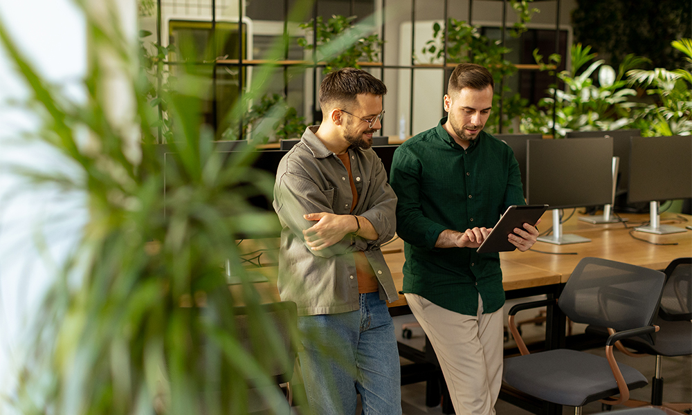 HR-Solutions_Kollegen-im-Office-mit-Pflanzen 2 Männer unterhalten sich im Büro und schauen auf ein Tablet. Das Büro ist begrünt.