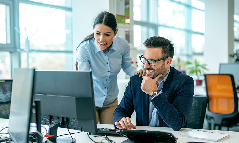 HR-Solutions_Kollegen-vor-Desktop Kollegen schauen gemeinsam auf einen Desktop auf dem Schreibtisch und unterhalten sich fröhlich