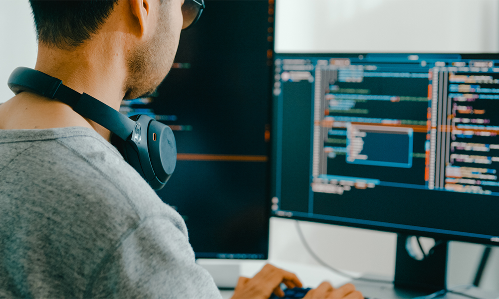 HR-Solutions_Mann-mit-Headset-vor-Bildschirm-Programmierer Programmierer sitzt vor einem Bildschirm und hat Kopfhörer um den Hals hängen